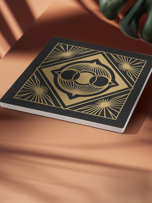 A black and gold ceramic coaster featuring a triple moon design framed by art deco geometric details and celestial sunburst accents.